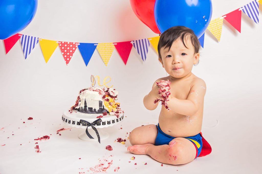 baby boy sitting next to a messy cake with frosting all over his hands and smiling at the camera during a superman cake smash photoshoot.