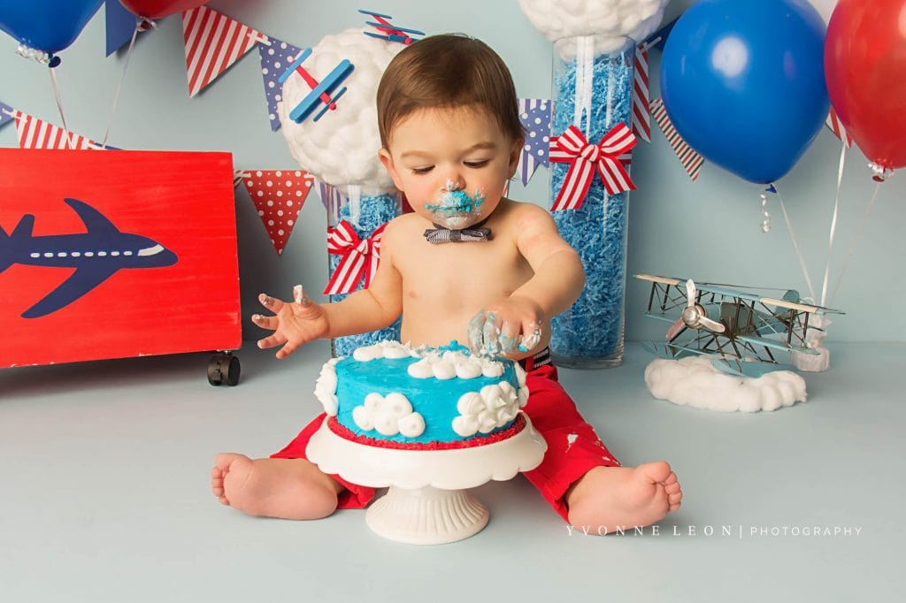 baby boy eating cake during an airplane theme cake smash photo shoot, cake is blue with white clouds made of frosting