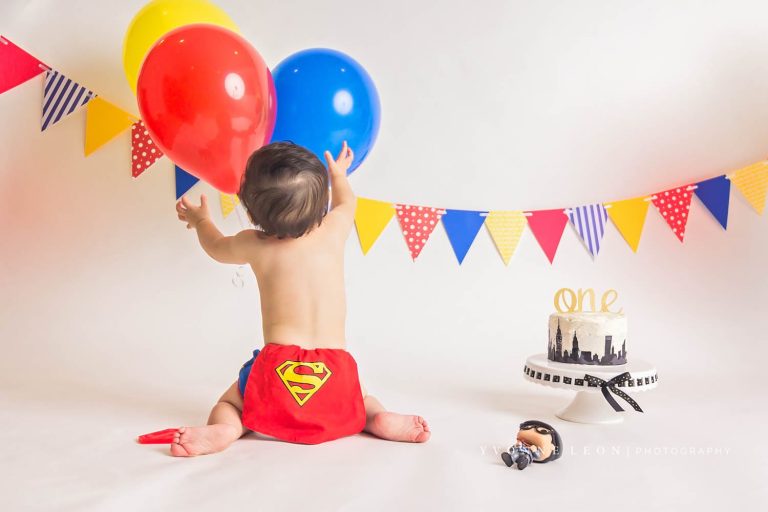 Baby boy wearing a diaper cover with a little cape and Superman logo for a Superman cake smash