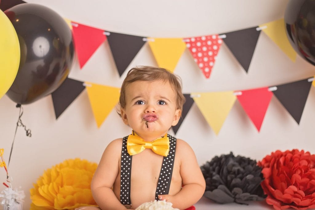 baby boy puckering his lips and making a duck face during his Mickey Mouse cake smash themed photoshoot; he wears a yellow bowtie and black suspenders