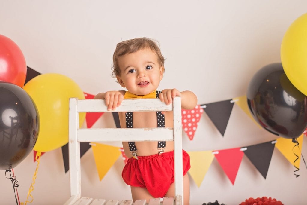 baby boy smiling at the camera as he peeks above the slats of a small white wooden chair he is holding onto