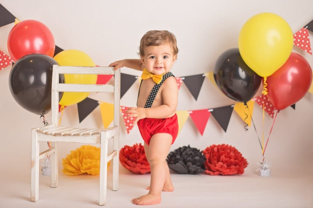 baby boy holding onto a chair as he stands sideways toward the camera and smiles during his cake smash photoshoot