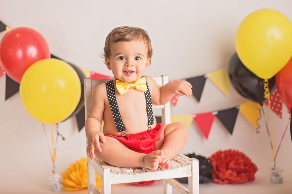 one year old boy sitting on a white chair smiling at the camera during his Mickey Mouse cake smash photo session