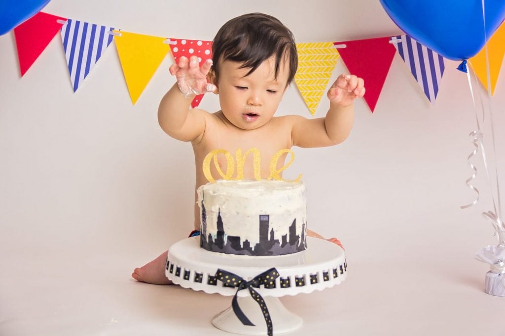 baby boy with hands up in the air looking like he is about to grab down on his birthday cake during a cake smash photoshoot.