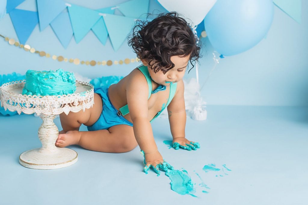 baby boy with blue frosting all over his hands and he is smearing it on the floor and leaving blue handprints