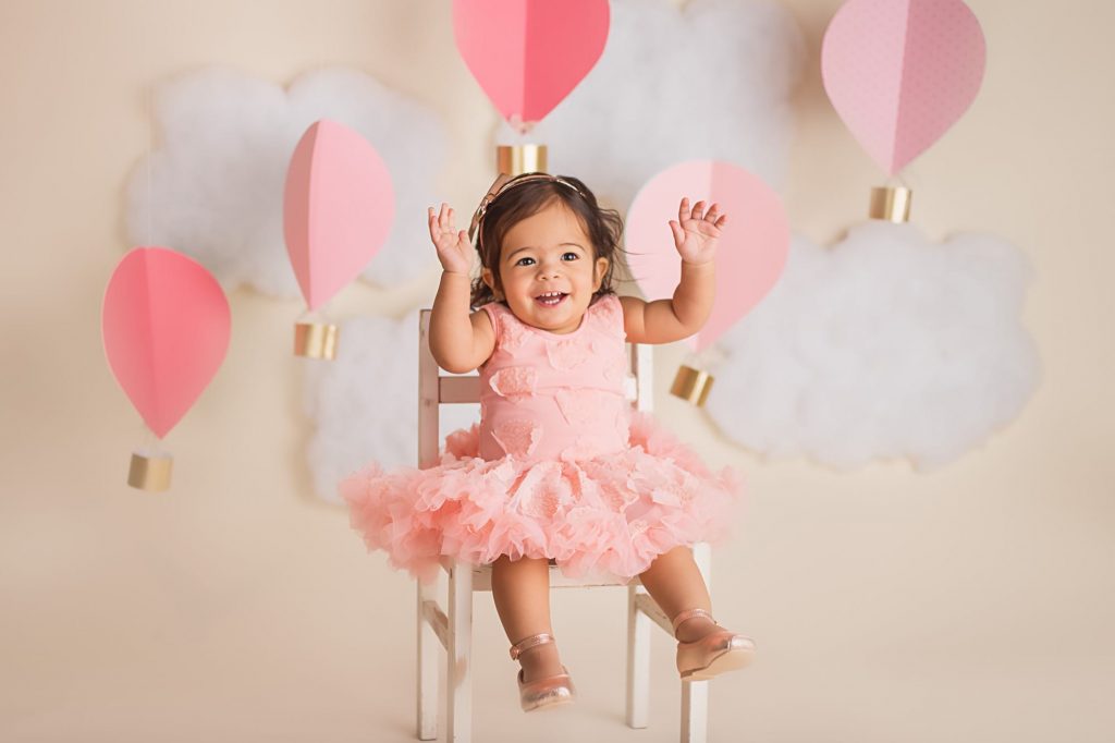 baby girl smiling and sitting on a chair for her hot air balloon cake smash