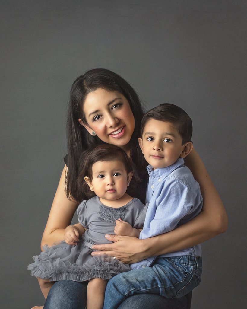 Photographer Yvonne Leon holds toddler daughter and preschool-aged son, both sit on her lap, all smiling; studio portrait.