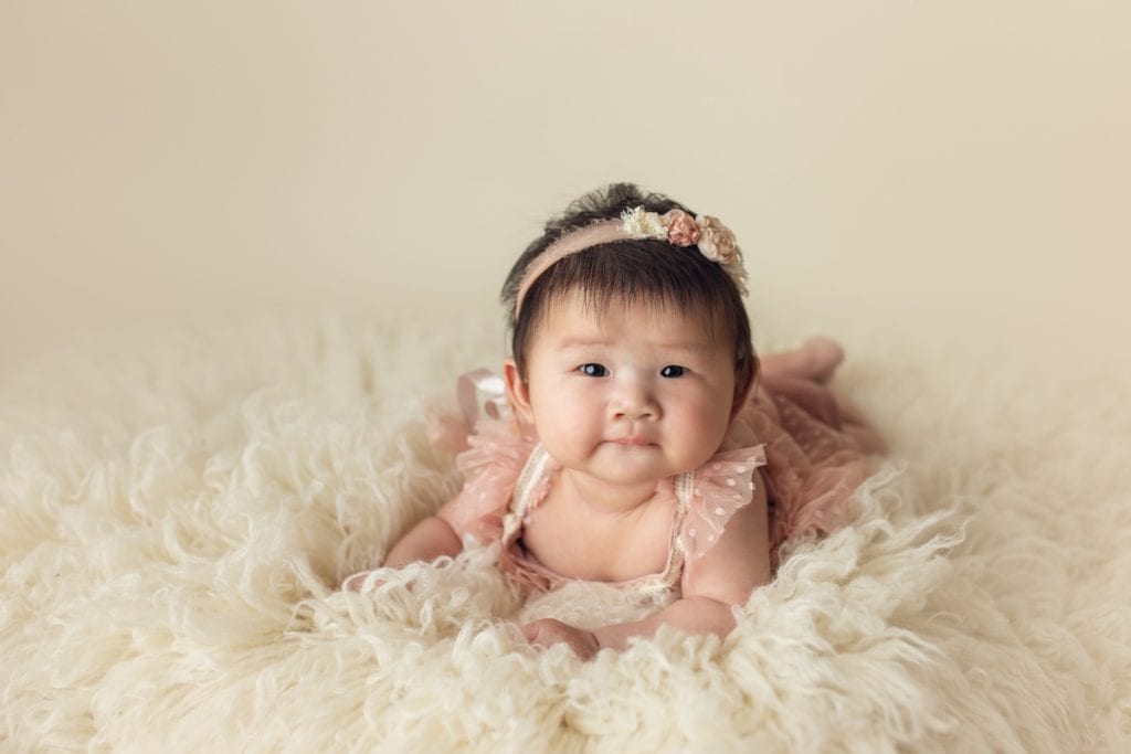 Infant girl in a pink dress laying on a fluffy cream-colored rug during a tummy time baby milestone studio photo session.