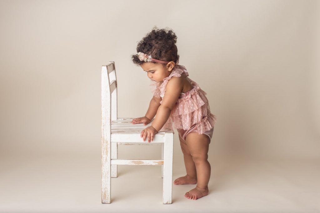 Baby girl in pink dress with chubby legs standing and holding onto a small chair prop during studio milestone photo session.