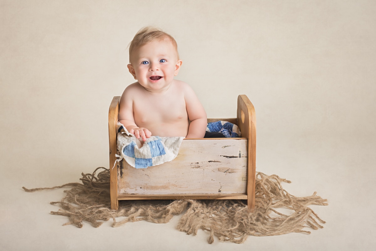 Laughing baby boy sitting in little bed prop with blue-and-white blanket sticking out the sides during milestone photoshoot.
