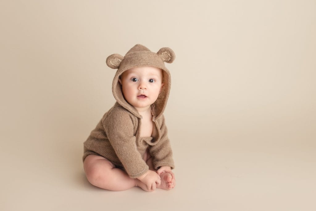 Smiling baby boy in a brown teddy bear hoodie sitting on a beige backdrop holding his toes during a studio photo session.