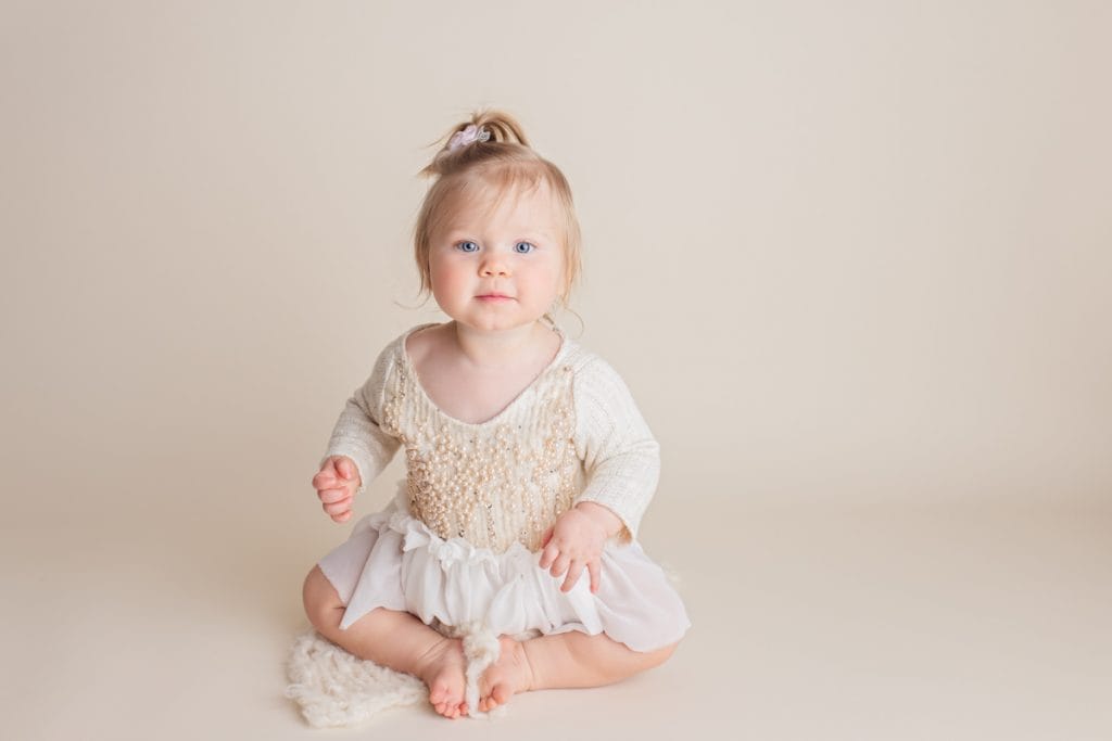 Smiling baby girl in a beaded white dress sitting up against a beige-colored backdrop during studio milestone photoshoot.