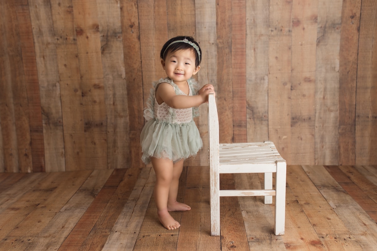 Baby girl standing while holding small white chair, wearing green dress, wooden plank backdrop during milestone session.