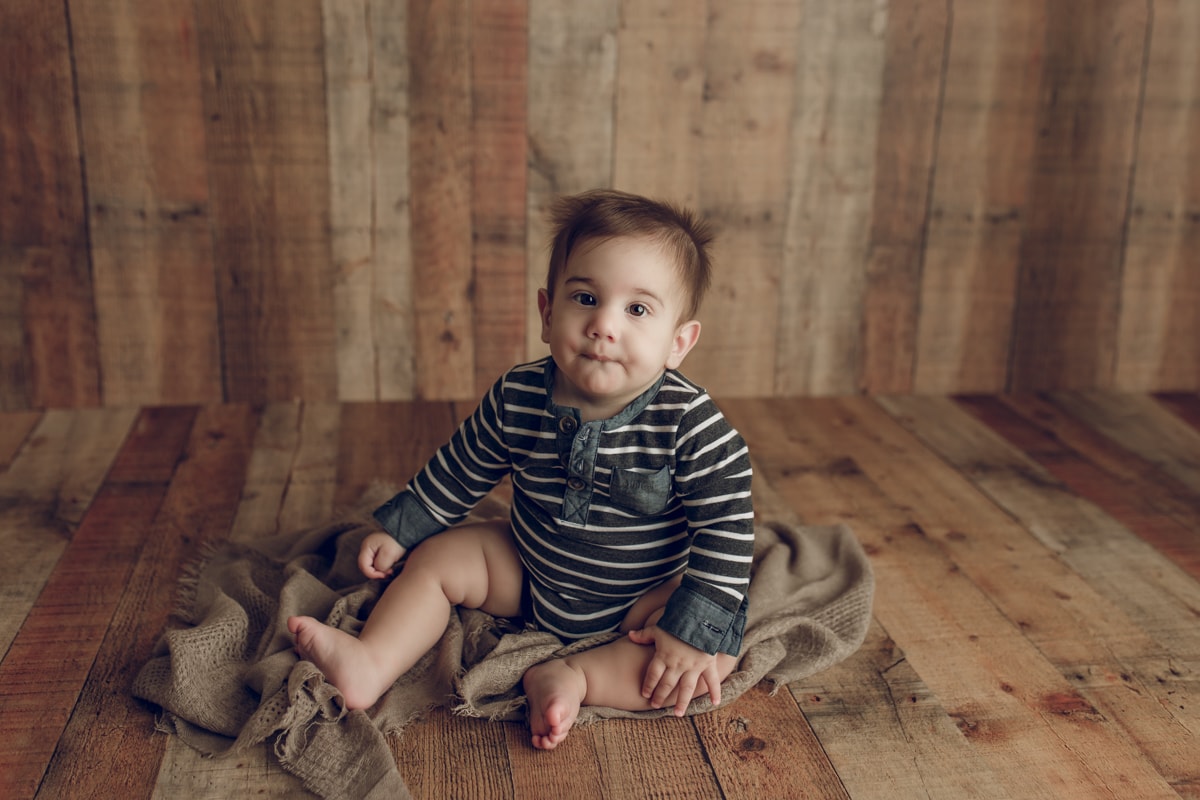 Baby boy in striped blue romper sitting and gazing at camera on a wooden panel backdrop during baby milestone studio session.