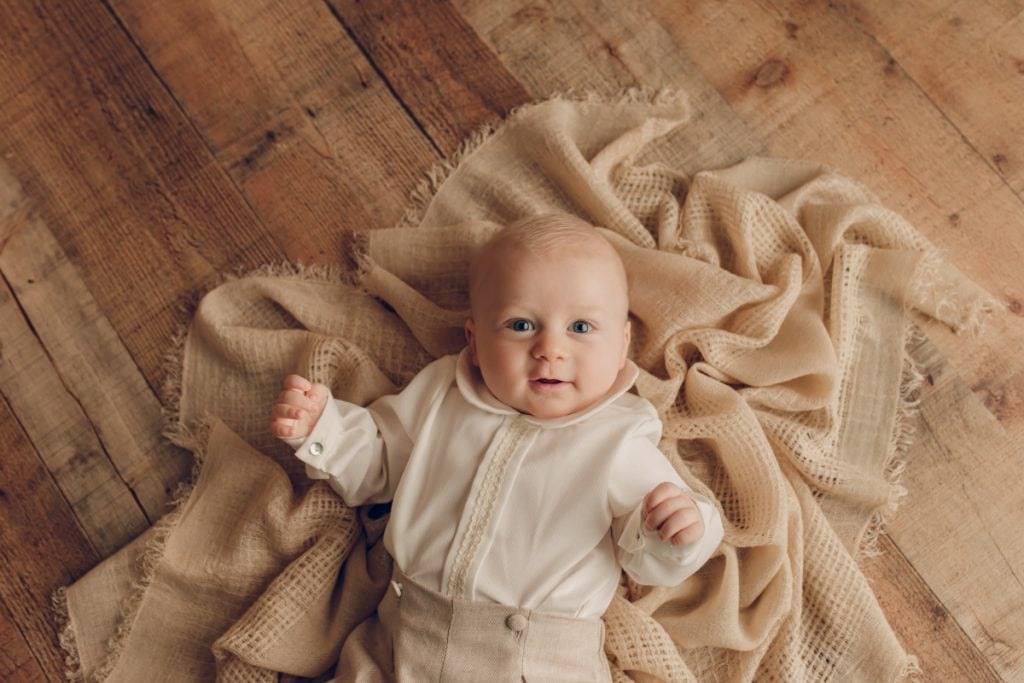 Baby boy wearing cute baby clothes laying on top of a blanket and looking up at the camera.