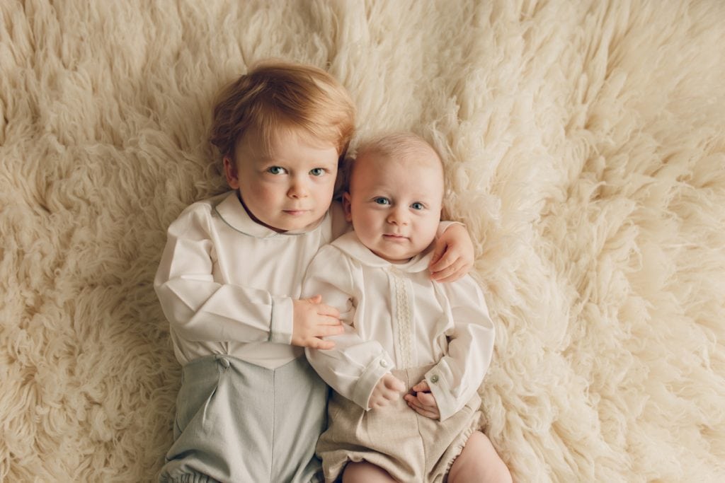Toddler and infant brothers in matching outfits lying on a fluffy rug looking up at camera during a milestone photo session.