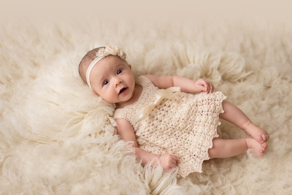Smiling baby girl in cream-colored crochet dress laying on fluffy rug and looking up at the camera during studio photoshoot.