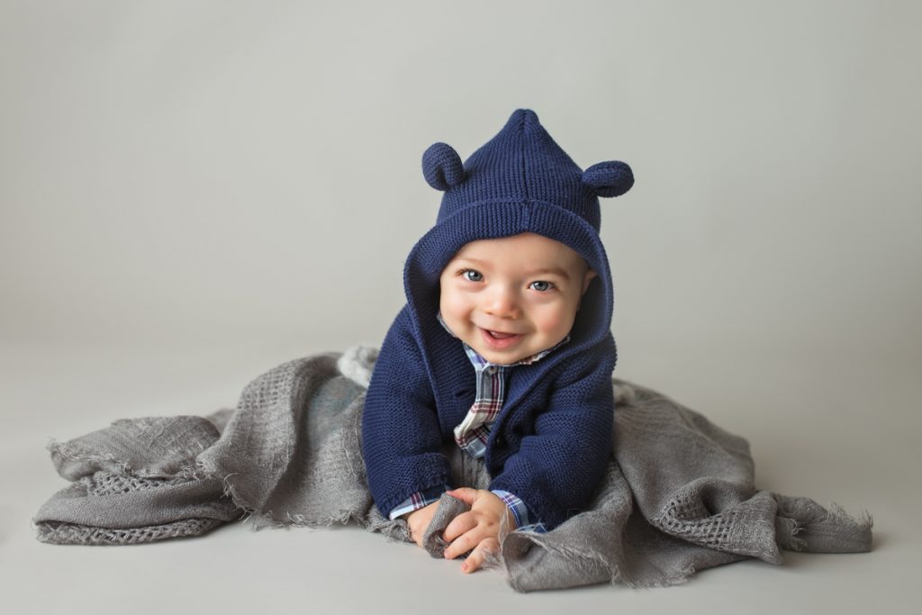 smiling baby boy in a blue teddy bear hoodie doing tummy time in a studio portrait.
