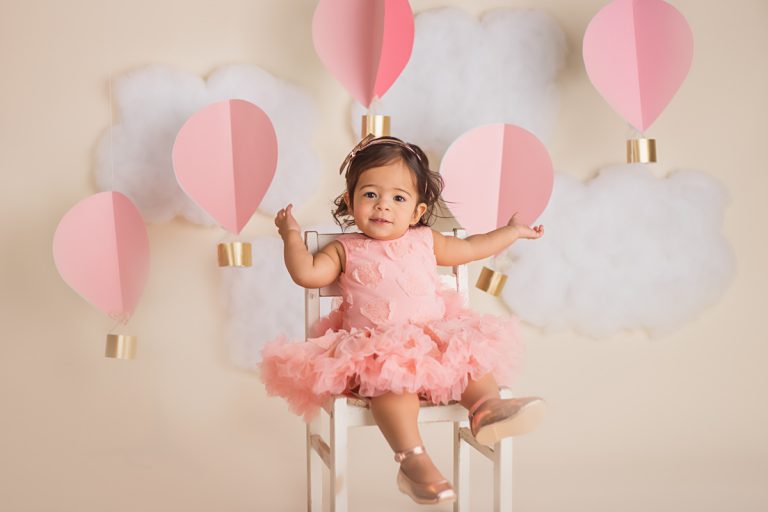 Smiling baby girl in pink dress during cake smash session with clouds and pink and gold paper hot air balloons in backdrop.