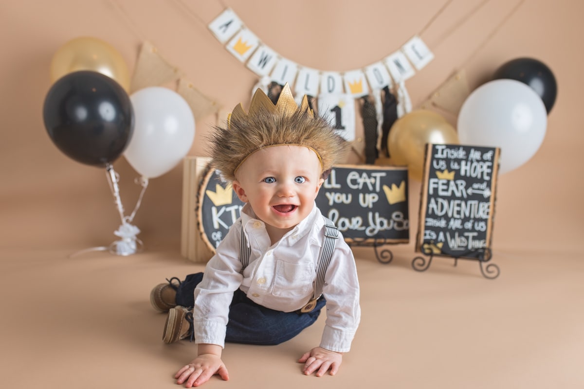 Smiling baby boy in suspenders and a furry crown during a "Where The Wild Things Are" themed cake smash photo session.
