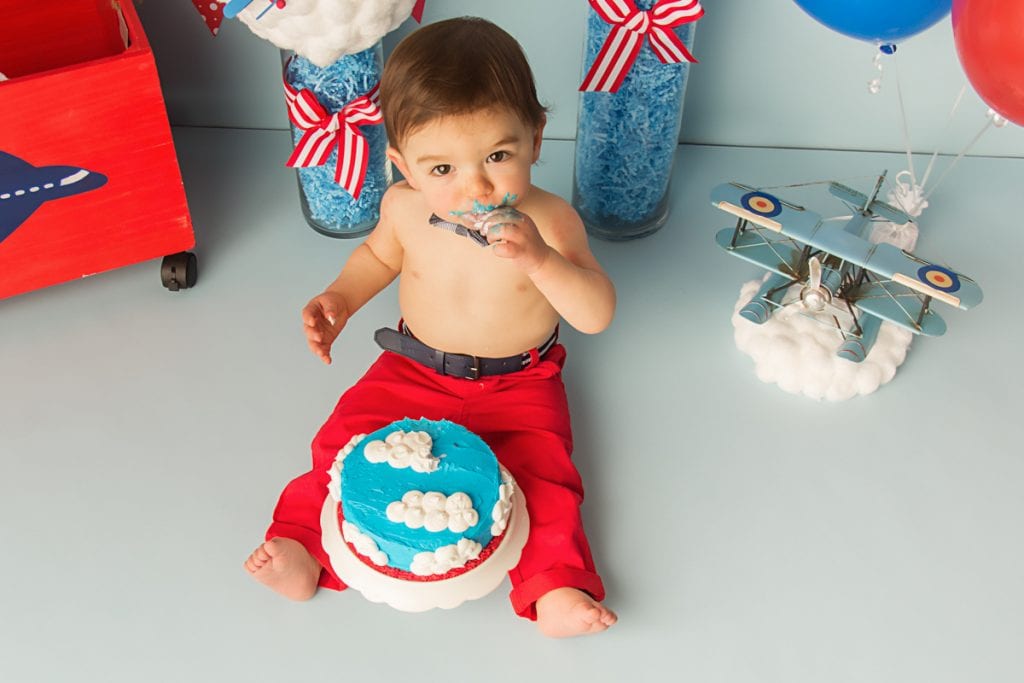 baby boy sitting in front of a birthday cake that is blue with white cloud frosting. He is taking a taste of his cake during his cake smash photoshoot for first birthday.