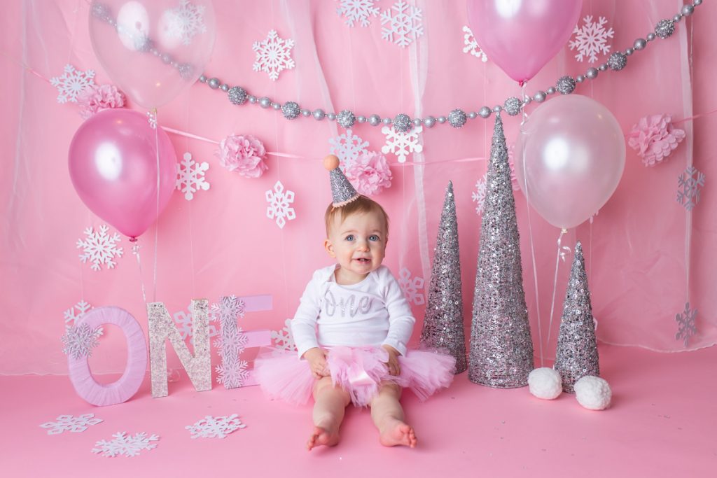 baby girl wearing white onesie with word "one" on front, pink tutu skirt and sparkly silver hat. Waiting for her cake during a winter onederland cake smash photoshoot.