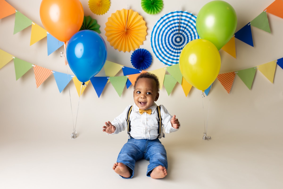 Baby boy in jeans and suspenders smiling during cake smash; festive backdrop of orange, blue, yellow, and green decorations.