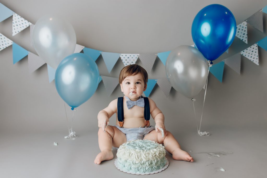 Baby boy in blue suspenders and bowtie sitting with cake during blue-and-grey cake smash; banner and balloons in background.