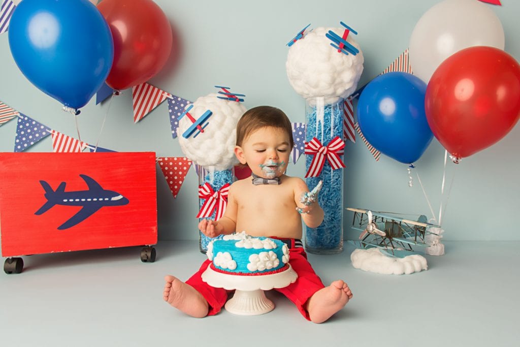 baby boy with blue frosting on his face after tasting his birthday cake and smiling while looking at the cake.
