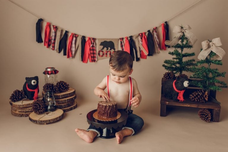 Baby boy in jeans and red suspenders about to dig into cake during rustic red-and-black plaid winter-themed cake smash shoot.