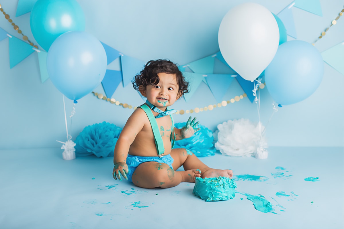 Smiling baby boy during a cake smash studio session with a blue backdrop and cake, frosting on his fingers and face.