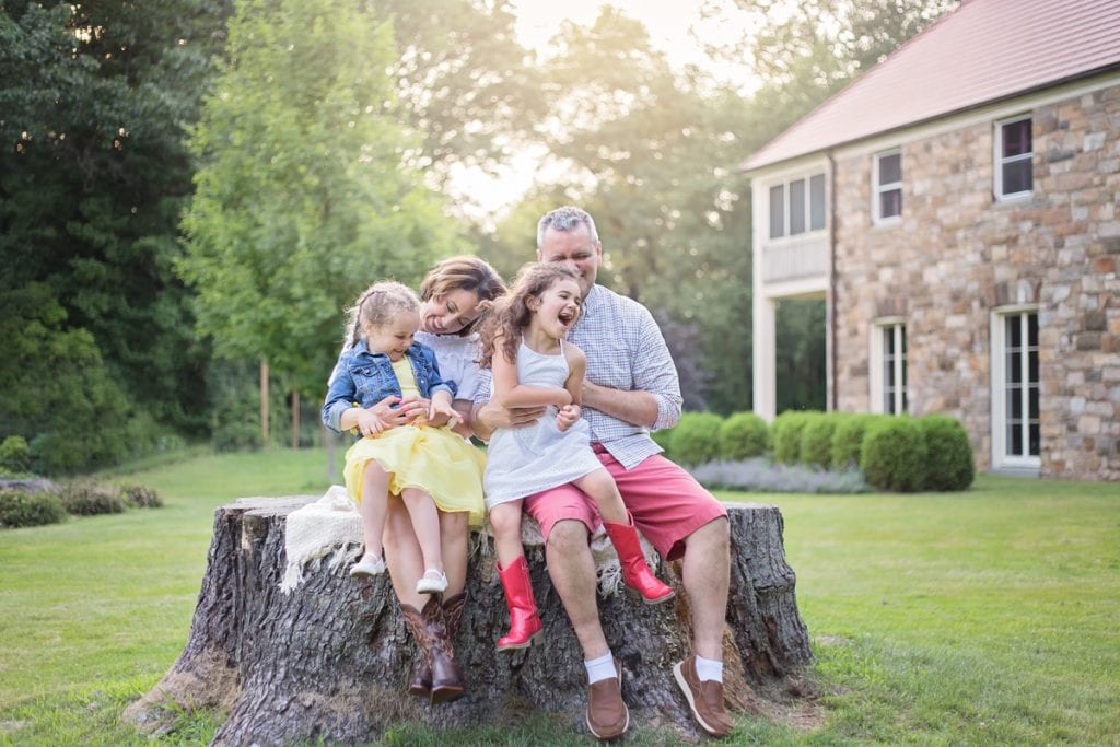 Family of four with mom, dad and two school-aged daughters laughing together on large tree stump; spring greenery background.