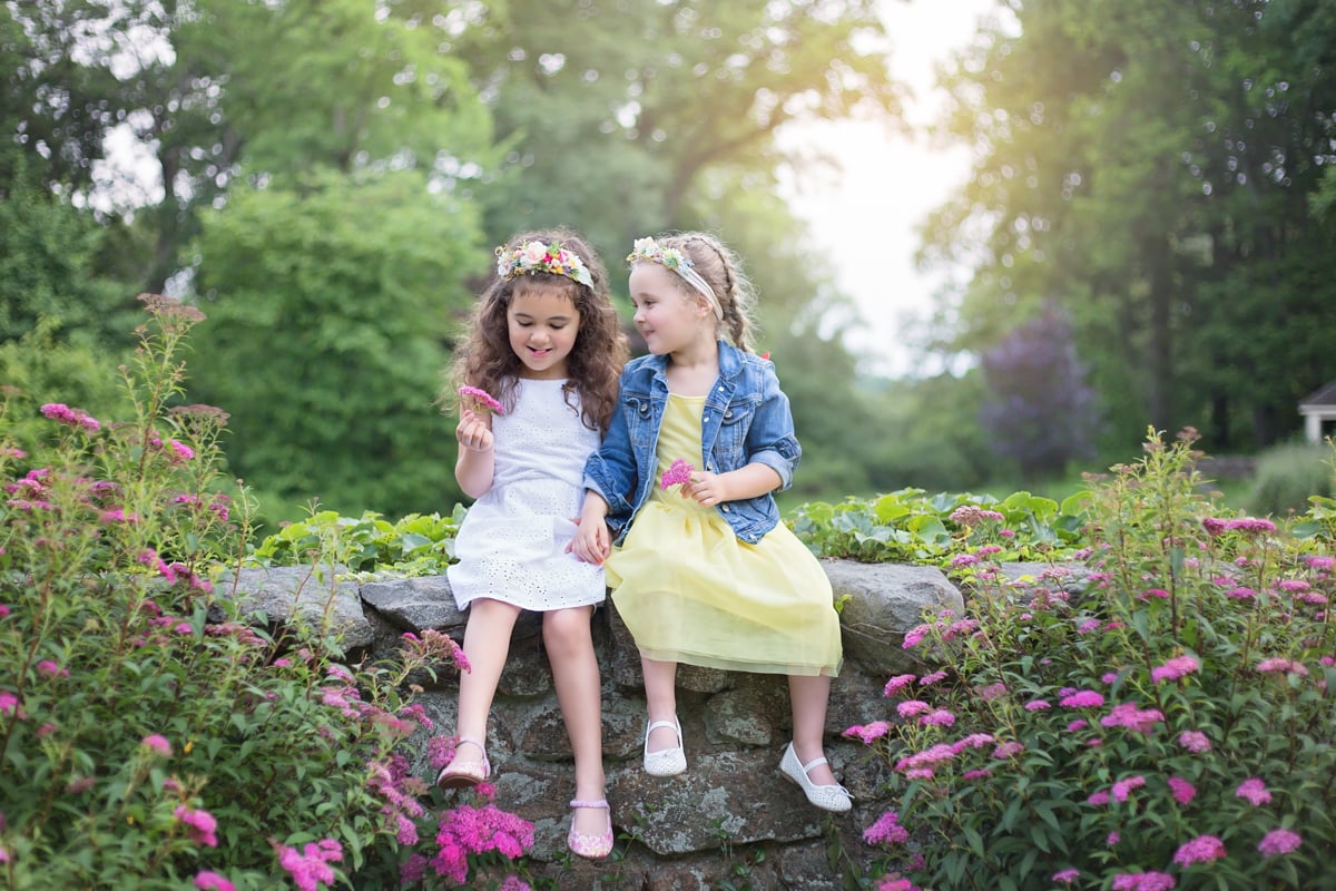 Two sisters in dresses and floral headbands giggling on rock wall surrounded by flowers during spring outdoor photo session.