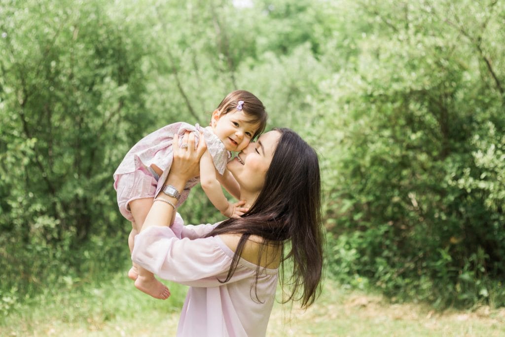 Mom snuggles toddler daughter who smiles at the camera; both wearing pastel pink outfits, soft spring greenery in background.