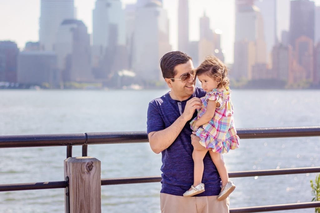 Dad holds toddler daughter by the waterfront on sunny day, NYC skyscrapers in the background; outdoor summer photo session.