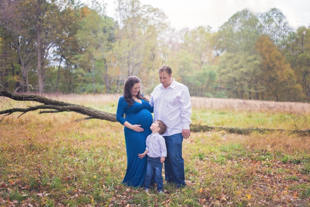 Family of three with mom, dad, and little boy all smiling at each other during an outdoor maternity photo shoot.  Little boy looks up at mom's belly.