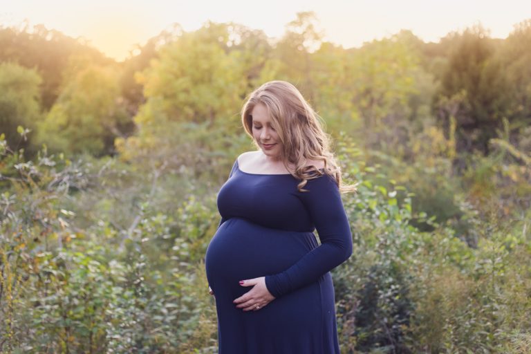 Pregnant mom in navy blue maternity dress holding and looking down at belly during outdoor sunset maternity session.