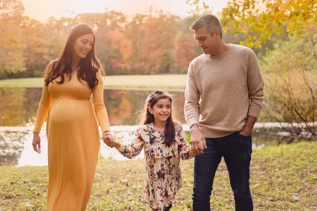 Fall family maternity session with school-aged daughter standing between parents holding hands, smiling, foliage behind.