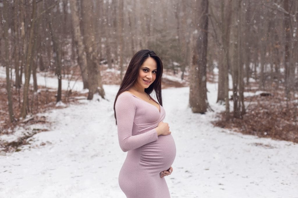 Mom-to-be cradling pregnant belly and smiling at the camera during a winter maternity portrait session in the snow.