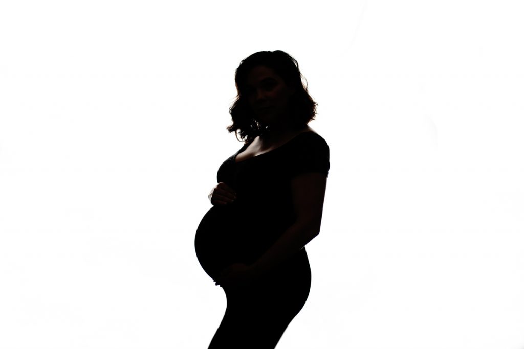 Backlit silhouette maternity portrait; dramatic lighting; subject appears blackened against white background, high contrast.