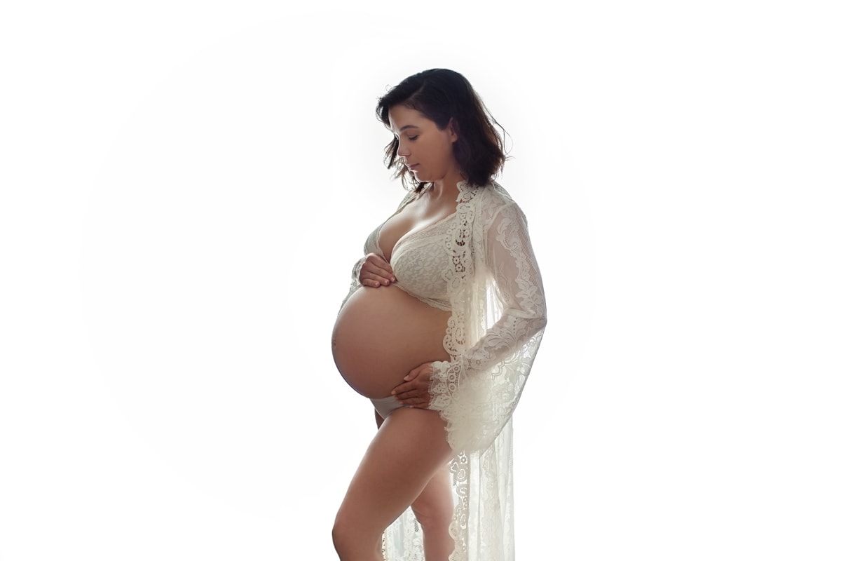 Backlit maternity studio portrait of mom in white lace bra and gown with exposed belly, glowing effect from light behind her.