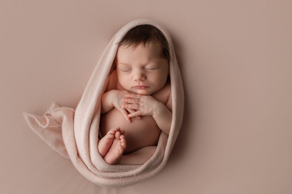 Newborn girl in egg wrap pose on a pink backdrop with feet together and hands flat on chest during a studio newborn session.