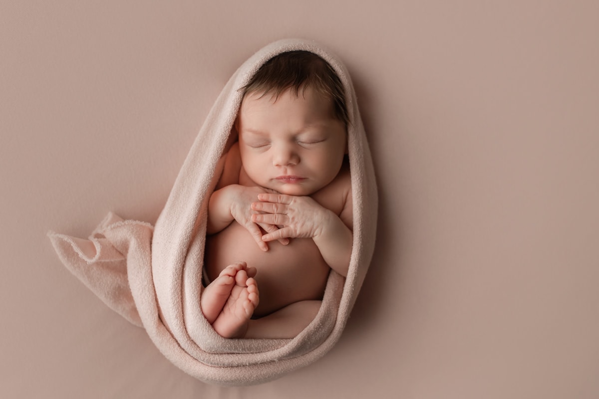 Newborn girl in egg wrap pose on a pink backdrop with feet together and hands flat on chest during a studio newborn session.