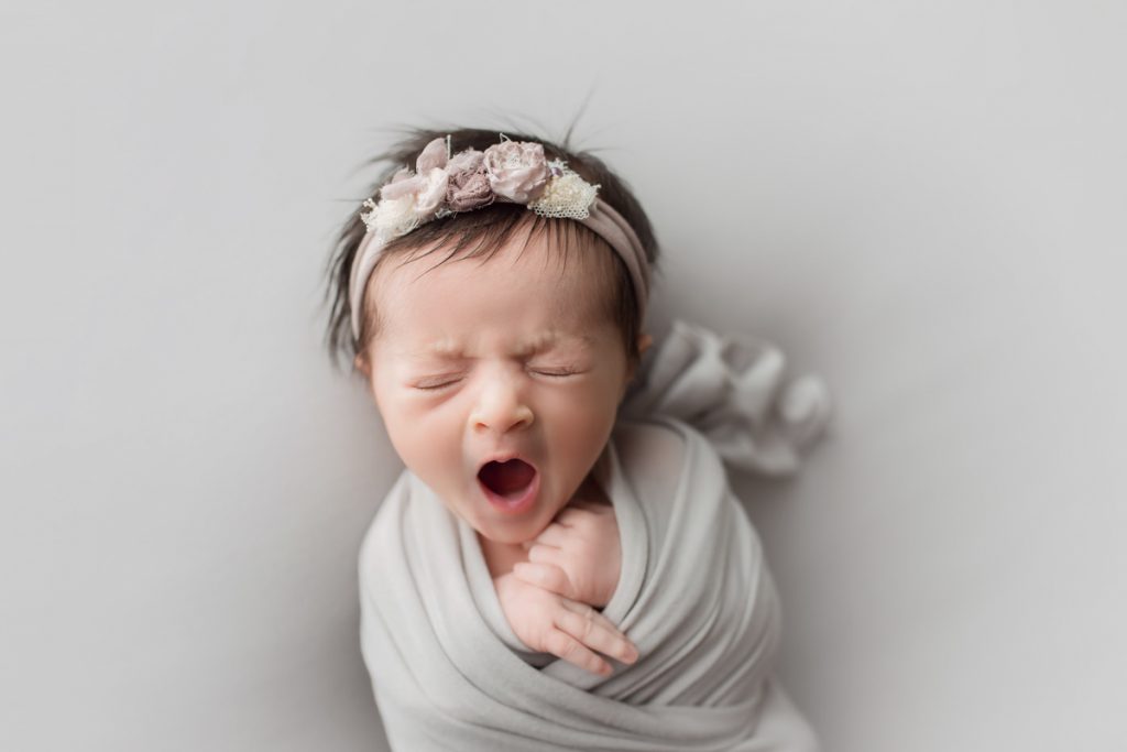 yawning newborn baby girl wrapped in grey and wearing a matching grey headband with bows and flowers.
