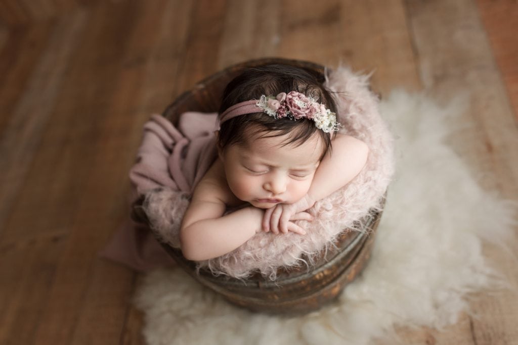 newborn baby girl sleeping in a bucket photography prop lined with a fuzzy pink blanket.