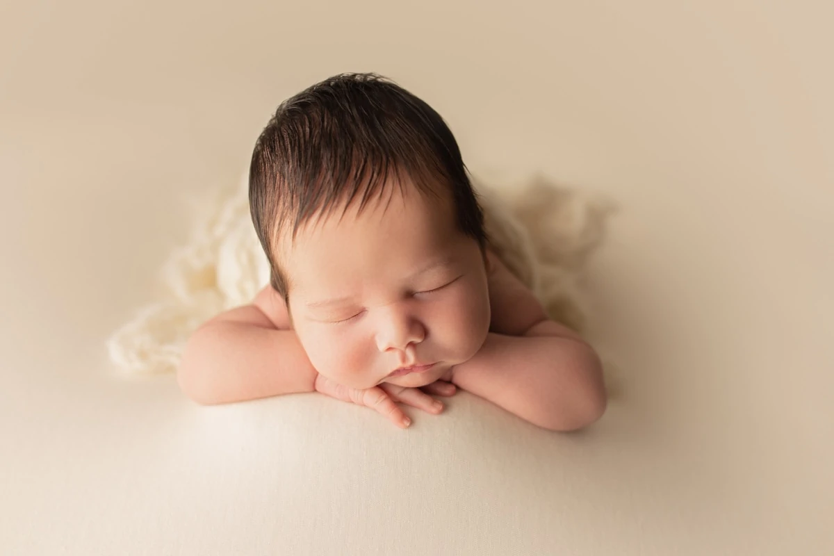 Sleeping newborn baby boy in chin-on-hands pose on a cream-colored backdrop during a studio newborn portrait session.