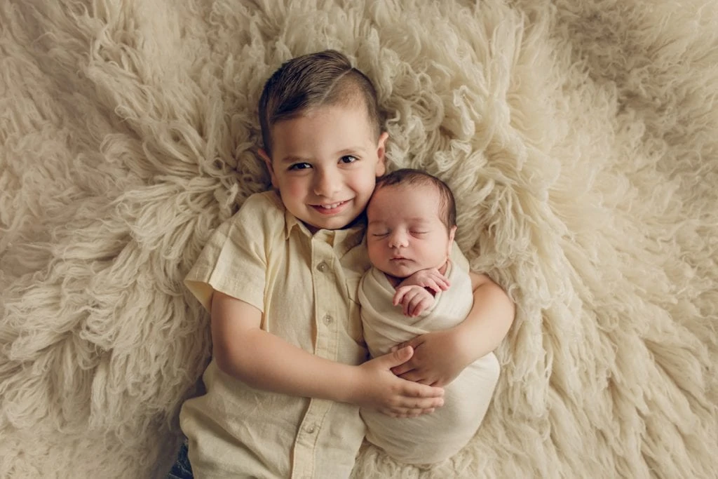Young boy hugs newborn brother on fluffy rug and smiles at camera while baby sleeps peacefully during studio newborn session.