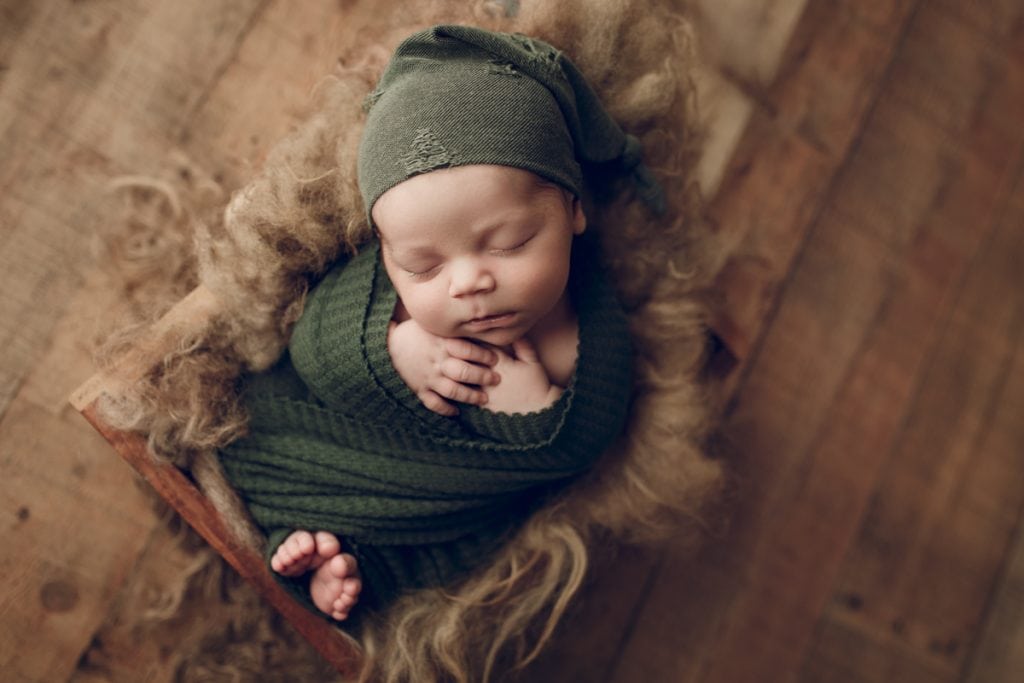 newborn baby boy wearing a green sleepy cap and nestled in a wooden crate lined with fur; newborn photography props.