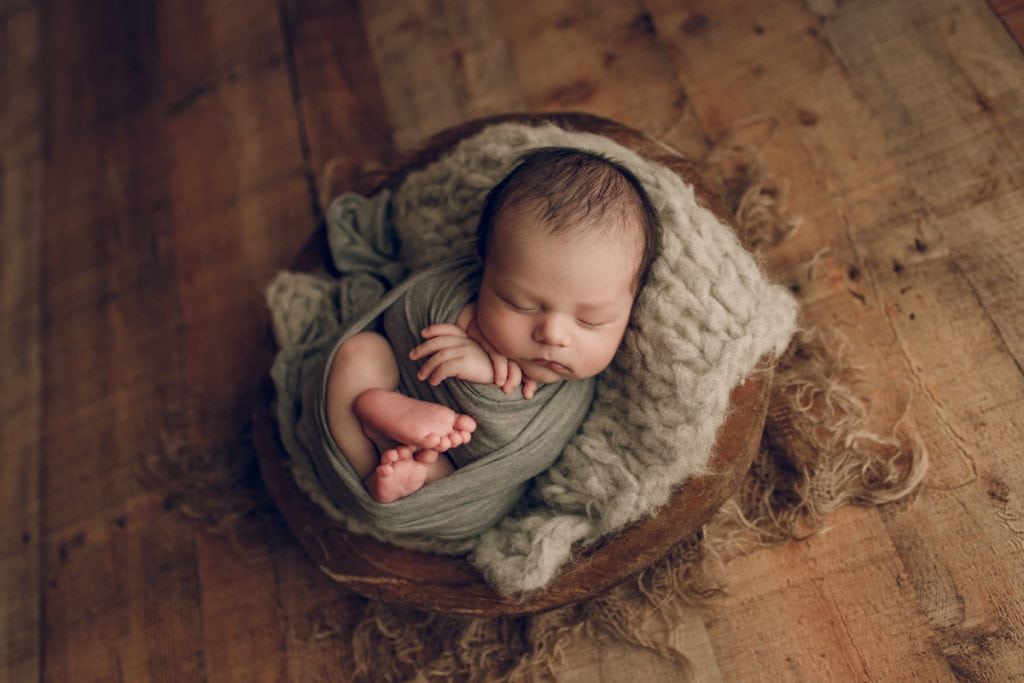 newborn baby boy curled up in a wooden bowl photography prop for a newborn portrait session.