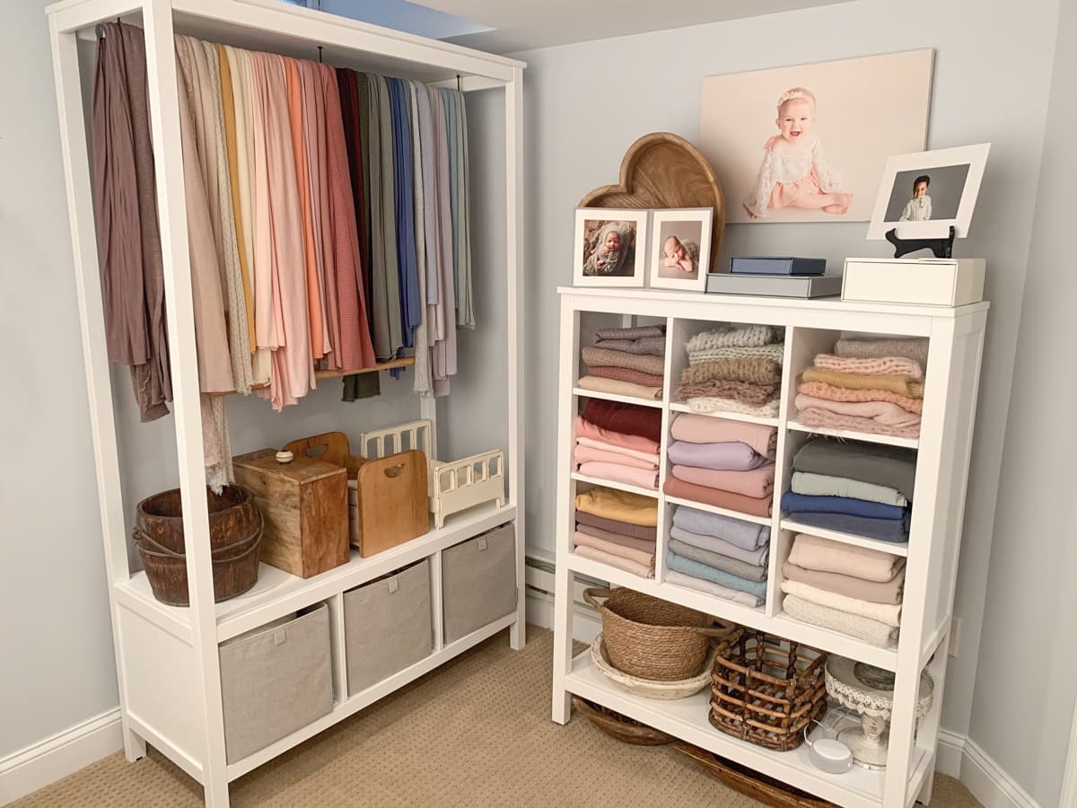 Corner of newborn photo studio with rack of wraps, folded backdrops organized by color, and floor props on display.